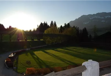 A tranquil landscape with a grassy meadow and long shadows in the sunlight. In the background, mountains and trees can be seen.