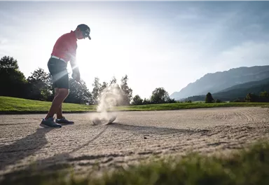 A golfer hits a ball out of the sand bunker. In the background, trees and mountains can be seen.