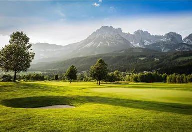 A picturesque golf course with lush green and gentle hills. In the background, majestic mountains rise under a clear sky.