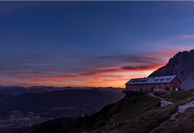 Ein malerisches Berghaus auf einem Hügel bei Sonnenuntergang. Der Himmel ist in warmen Farben gefärbt und die umliegenden Berge sind deutlich sichtbar.