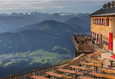 Ein Bergrestaurant mit einer Terrasse und Sitzplätzen im Freien. Im Hintergrund sind majestätische Berge und eine grüne Landschaft zu sehen.