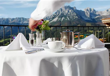 Ein gedeckter Tisch mit einer Teekanne und einer Blumenvase in den Bergen. Die Umgebung ist malerisch mit klarer Aussicht auf die Berge.