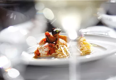 Ein elegantes Gericht auf einem weißen Teller, dekoriert mit frischen Zutaten. Im Hintergrund sind unscharfe Gläser sichtbar, die eine gehobene Atmosphäre schaffen.