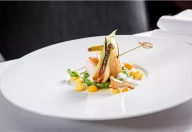 Ein kunstvoll angerichtetes Gericht auf einem weißen Teller. Es enthält Fisch, Gemüse und farbige Saucen als Garnierung.