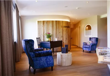 Ein eleganter Empfangsbereich mit blauen Sesseln und einem runden Holztisch. Im Hintergrund befindet sich eine Holzverkleidung mit Pflanzen.