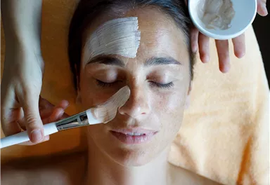 Eine Frau liegt entspannt auf einer Liege, während ihr Gesicht mit einer Gesichtsmaske behandelt wird. Eine Hand hält einen Pinsel, um die Maske aufzutragen, während die andere Hand eine Schüssel mit produktivem Inhalt hält.