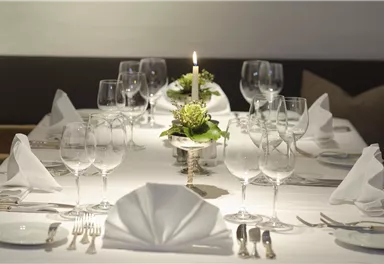 Ein elegant gedeckter Tisch mit Gläsern, Besteck und Servietten. In der Mitte steht eine Kerze mit frischen Blumen.