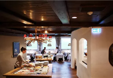 Ein gemütliches Restaurant mit rustikaler Holzdecke und elegantem Ambiente. Einige Gäste genießen ihre Mahlzeiten, während das Personal den Tisch vorbereitet.