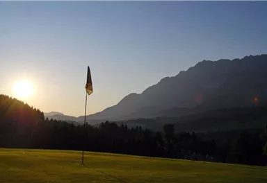 A quiet golf course with a flag, surrounded by mountains. The sun rises behind the hills, creating a peaceful atmosphere.