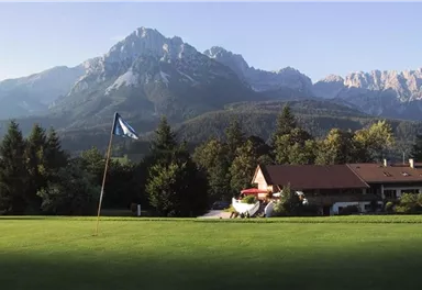 A beautiful golf course with a flagpole in the foreground. In the background, majestic mountains and a typical alpine house can be seen.