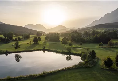 A picturesque landscape with gentle hills and a tranquil pond. The sun sets behind the mountains, casting warm light over the scene.