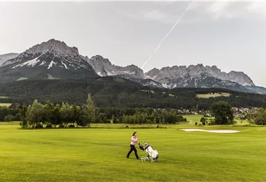 A person is driving a golf cart across a green golf course. Impressive mountains can be seen in the background.