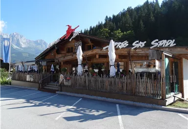 Ein traditionelles Restaurant in einer malerischen Umgebung mit Holzverkleidung und Sonnenschirmen. Im Hintergrund sind Bäume und Berge sichtbar.