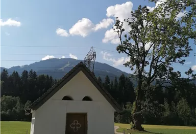 A small white chapel stands in a picturesque landscape. In the background, there are mountains and a blue sky with white clouds.