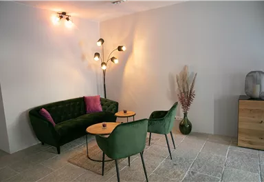 Ein stilvoll eingerichteter Raum mit einem grünen Sofa und zwei Stühlen. Auf einem kleinen Tisch stehen dekorative Elemente und die Beleuchtung schafft eine gemütliche Atmosphäre.