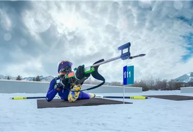 Ein Skisportler liegt im Schnee und zielt mit einem Gewehr auf eine Zielscheibe. Im Hintergrund sind schneebedeckte Berge und ein bewölkter Himmel zu sehen.