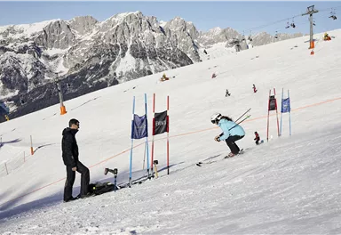 Eine Skifahrerin trainiert auf einer Piste in den Bergen. Im Hintergrund sind weitere Skifahrer und Lifte zu sehen.