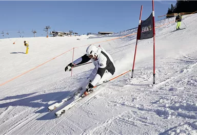 Ein Skifahrer fährt schnell durch einen Slalomparcours im Schnee. Im Hintergrund sind andere Skifahrer und Skilifte zu sehen.