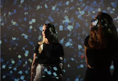 Two women are standing in front of a colorful, bright wall projection. The lights reflect on their faces and create a creative atmosphere.