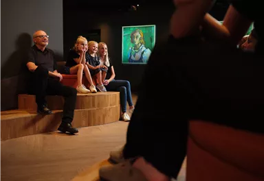 A group of people is sitting on a wooden bench in an art museum. In the background, a painting can be seen.