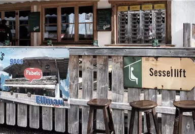 A rustic mountain cabin with a wooden bar and a sign for the chairlift. In the background, drinks and an inviting atmosphere can be seen.