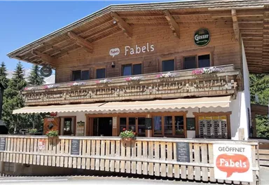 A beautiful wooden building with an inviting terrace. The restaurant "Fabels" has a rustic design and is surrounded by trees.