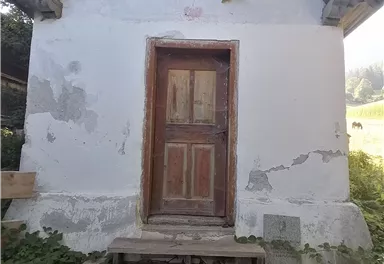 Ein kleines, einfaches Gebäude mit einer Holzfronttür. Es steht vor einer grünen Landschaft und ist von Gras und Pflanzen umgeben.