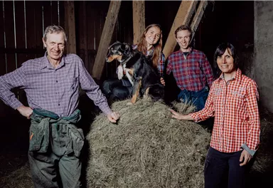 Eine Gruppe von vier Personen steht in einem Stall neben einem Heuhaufen. Ein Hund sitzt auf dem Heu und alle lächeln in die Kamera.