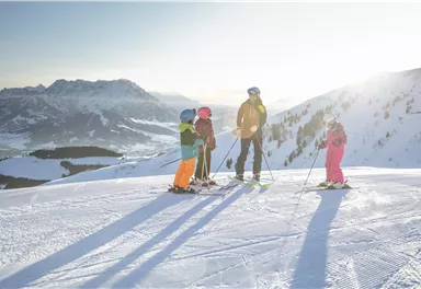 A group of children and an adult is standing on a snow-covered slope. The sun is shining and the mountains are visible in the background.