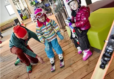 Two children are putting on ski boots while an adult helps them. In the background, ski equipment and a cozy room can be seen.