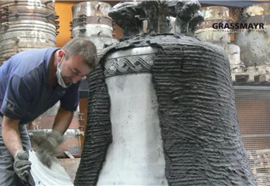 A craftsman is working on a large bell, which is being processed with a special mixture. In the background, more bells are visible.