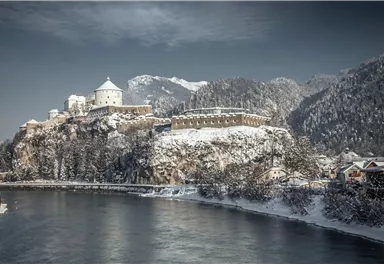An impressive castle on a snowy hill with mountains in the background. The river reflects the winter landscape.