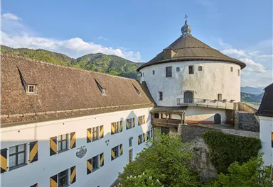 A historic building with a round tower and a spacious grounds. In the background, green hills and a blue sky are visible.