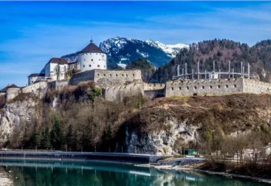 Eine historische Burg auf einem Hügel mit schneebedeckten Bergen im Hintergrund. Ein klarer Fluss fließt im Vordergrund.