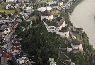 An impressive castle on a hill with surrounding houses and a river. The landscape is green and picturesque.