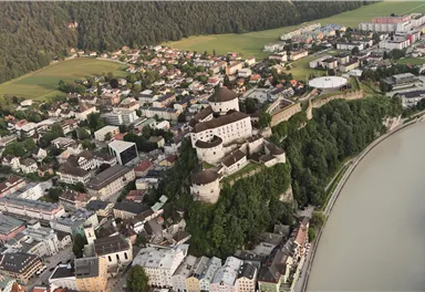 A picturesque view of a city with a historical castle on a hill. In the background, green meadows and the course of a river can be seen.