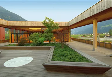 Ein moderner Innenhof mit Holzboden und umliegenden Pflanzen. Im Hintergrund sind Berge und ein klarer Himmel zu sehen.