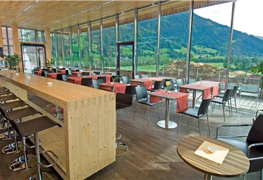 Ein modernes Restaurant mit großen Fenstern und Blick auf die Berge. Es gibt eine lange Holzbar und Tische mit roten Tischdecken.