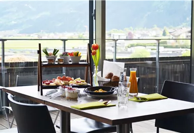 Ein eleganter Tisch mit Frühstücksspezialitäten und frischen Blumen. Im Hintergrund sind die Berge und eine grüne Landschaft sichtbar.