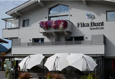 Ein charmantes Café mit dem Namen „Fika Bunt“ in einem modernen Gebäude. Im Vordergrund stehen Schatten spendende Sonnenschirme.