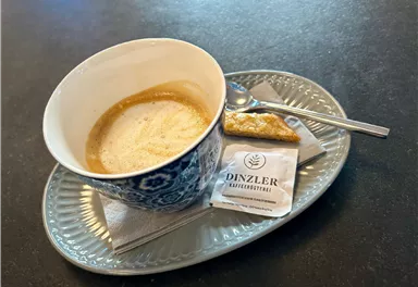 Eine Tasse Kaffee mit Crema auf einem eleganten Teller. Daneben befinden sich ein Gebäckstück und ein Zuckersüßstoff.