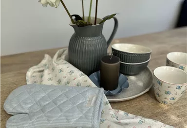 Eine stilvolle Tischdecko mit einem grauen Krug, weißen Blumen und Geschirr. Auf dem Tisch liegen ein Ofenhandschuh und ein Tuch.