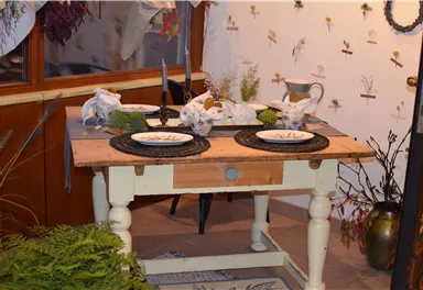 Ein schön dekorierter Tisch mit Tellern und Naturmaterialien. Im Hintergrund sind Blumen und Pflanzenmuster an der Wand zu sehen.