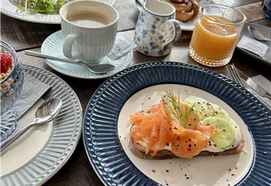 Ein reichhaltiges Frühstück mit einem offenen Sandwich mit Räucherlachs und Gurken. Dazu Kaffee, Saft und einige süße Köstlichkeiten auf dem Tisch.