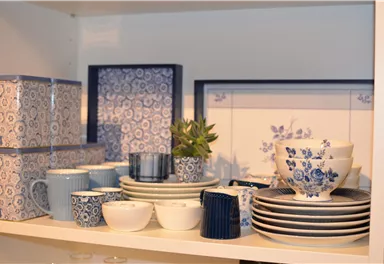 Eine Auswahl an blau-weißen Geschirr und Tassen steht auf einem Regal. Daneben sind dekorative Boxen und eine kleine Pflanze zu sehen.