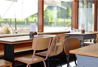 Ein einladendes Café mit Holzstühlen und Tischen. Große Fenster bieten einen Blick auf die grüne Umgebung.