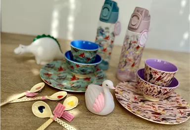 Eine farbenfrohe Kinder-Tafel mit Tellern, Tassen und Löffeln. Einige Flaschen und ein Spielzeug-Dinosaurier sind ebenfalls zu sehen.