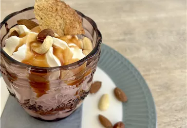 Ein Dessert in einem Glas mit Schlagsahne, Karamellsauce und Nüssen. Darum herum liegen einige Mandeln und Cashewkerne.