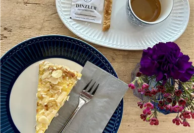 Ein schöner Teller mit einer Scheibe Kuchen, garniert mit Mandeln, und einer Gabel. Daneben befindet sich eine Tasse Kaffee und eine kleine Süßigkeit, umgeben von einer bunten Blumendekoration.