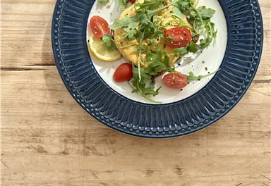 Ein appetitlicher Teller mit gebratenem Fisch, garniert mit frischem Rucola, Cherrytomaten und Zitronenscheiben. Der Teller liegt auf einem rustikalen Holztisch.